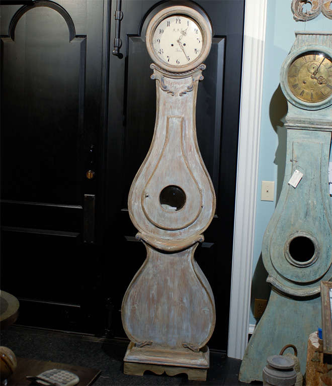This 19th century Swedish clock is of a grey or brown color with blue painted accent. It has a perfectly round head supported by a beaded neck sitting on a voluted collar. In the middle is a tear shaped door centered on a convex glass window. Below