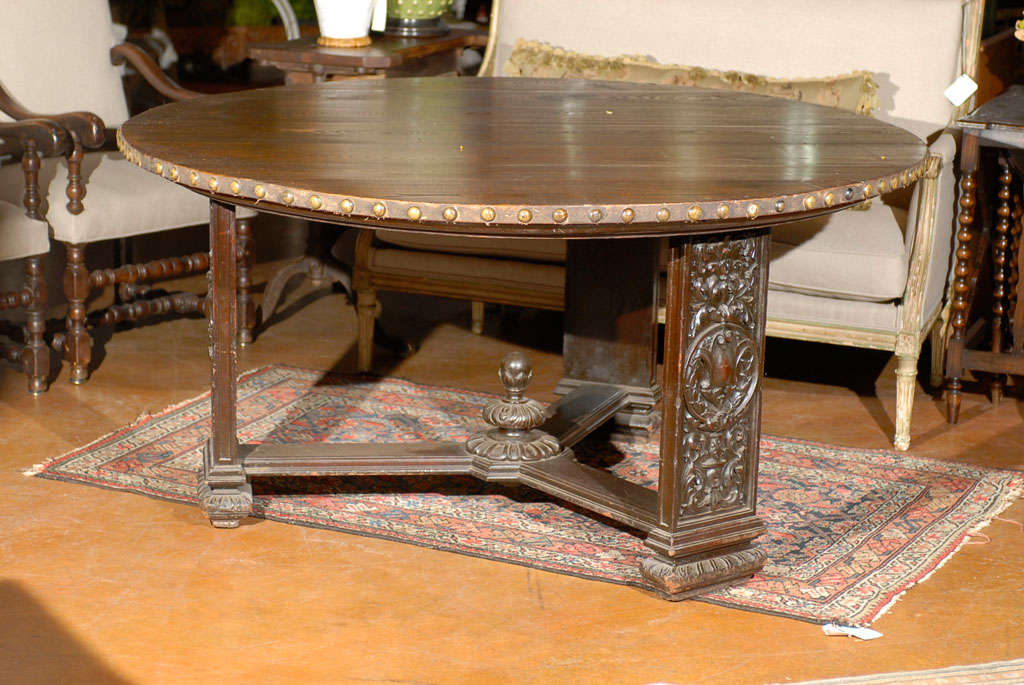 French circular oak table, the top surmounting a studded metal apron raised on three flattened rectangular carved legs and joined by a Y-form stretcher terminating in a central finial. 