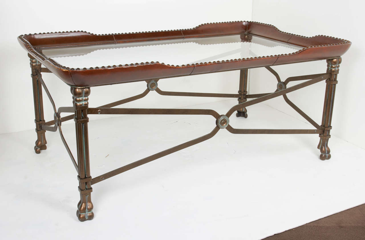 Handsome coffee table with patinated bronze base and a glass top center. The tabletop has an elegant sinuous design comprised of leather wrapped wood in a beautiful cognac color with studded bronze details. The base features stylized cross bars