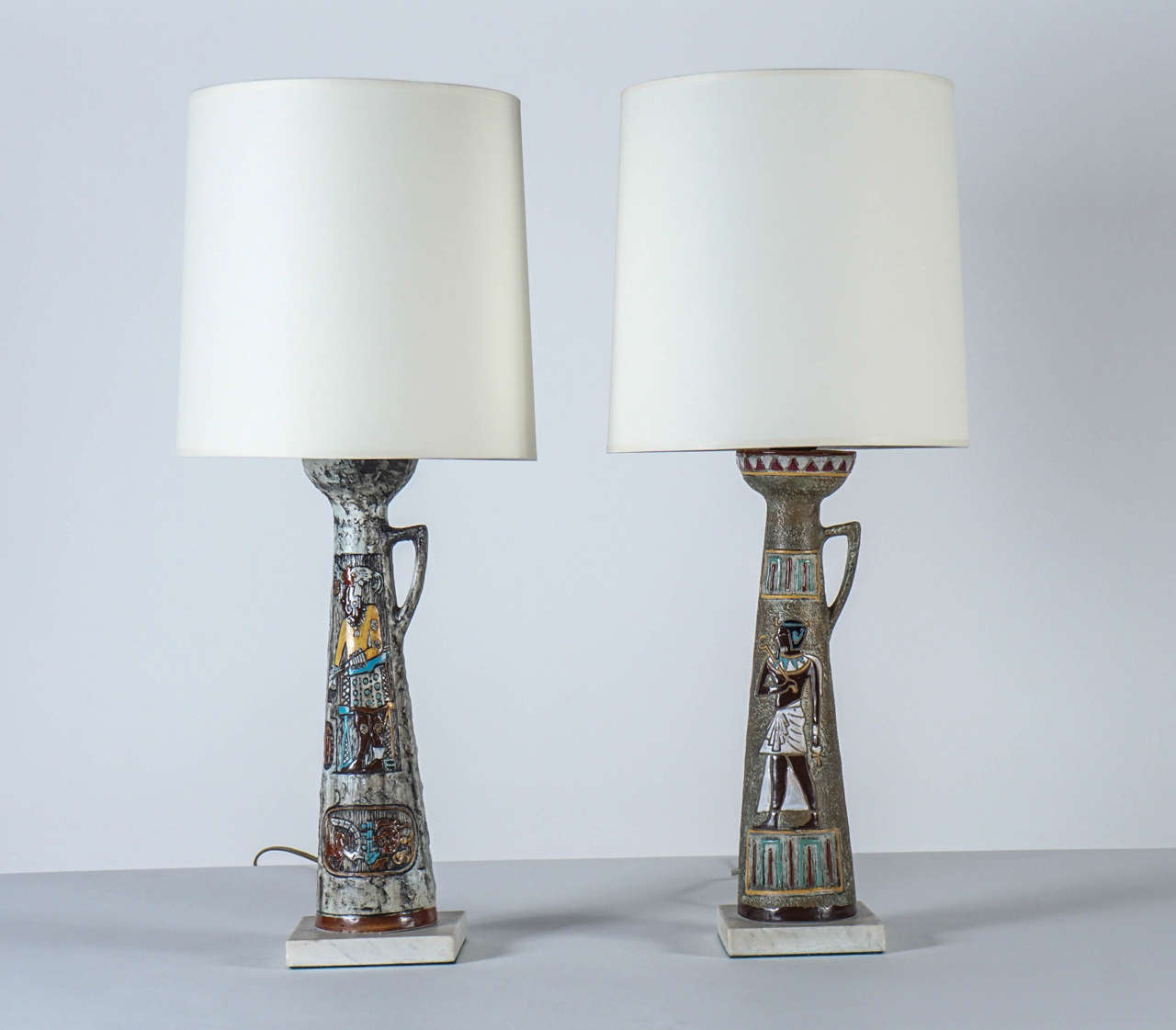 Egyptian-motif ewer-form bases: different hieroglyphic figures are depicted on each lamp in chocolate, mustard, and turquoise against a dove gray lava-glaze ground. White marble bases. Newly rewired with 3-way sockets. Ivory paper shades included.