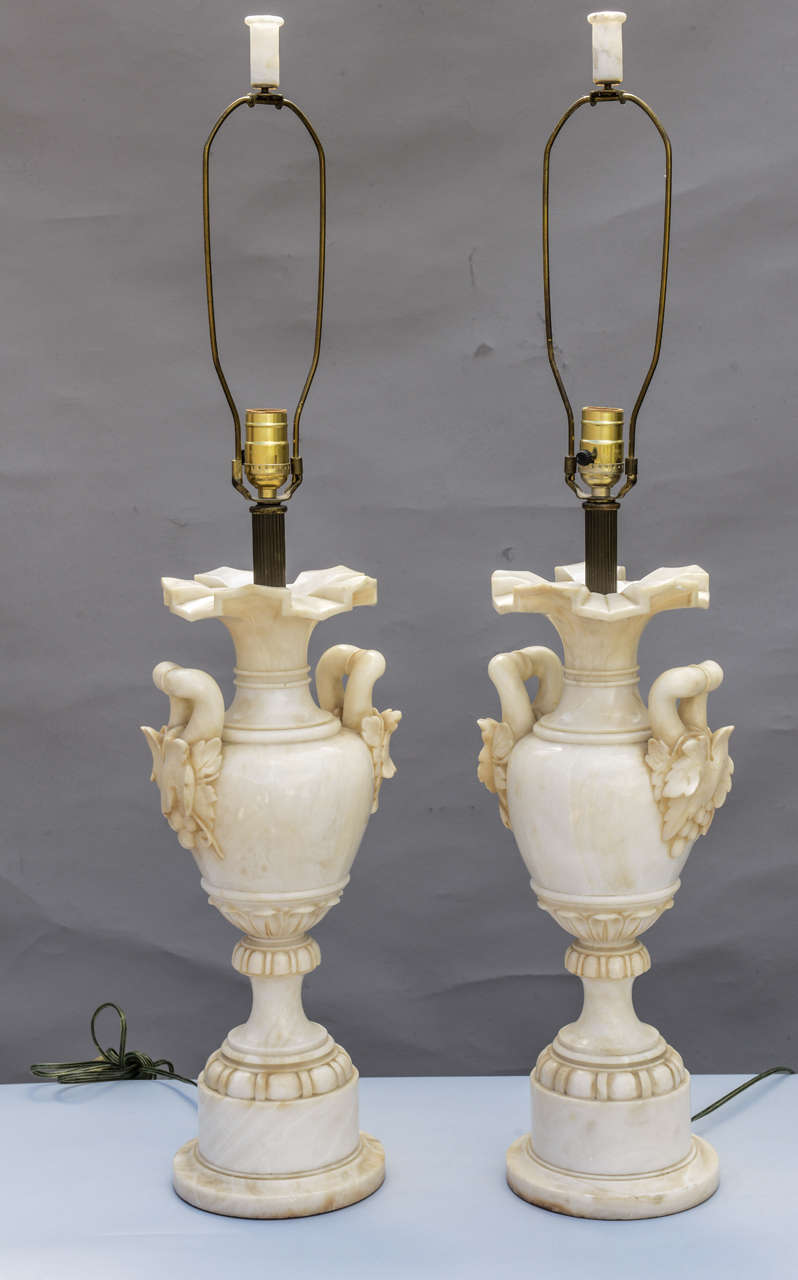 Pair of lamps, each a carved urn of alabaster with fine patina, large handles decorated with grapes, neck finished with geometric flared lip, raised on round foot on conforming foot. Matching finials.