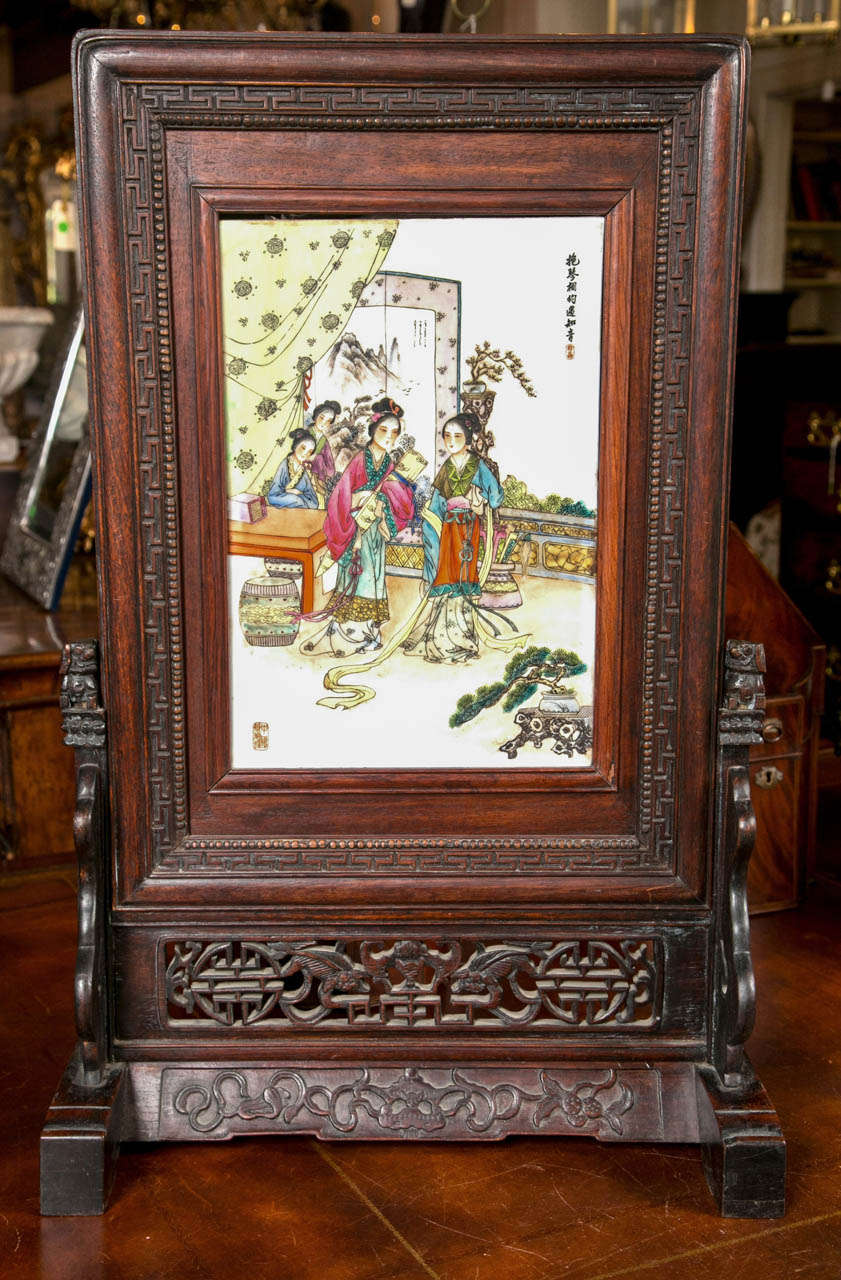 Beautifully carved rosewood frame, stands on its own .  The porcelain plaque depicts  women in an outdoor room setting with a screen and drapery behind them. A low  red table, a  garden seat and a potted bonsai tree on a tree trunk stand complete