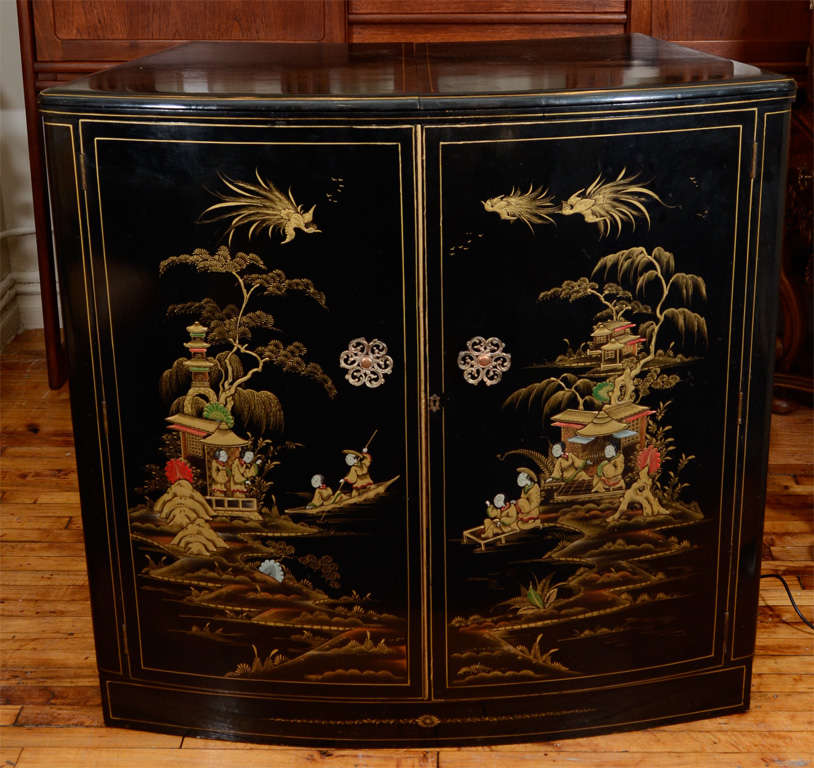 An English art deco cocktail cabinet or bar in ebonized wood with chinoiserie landscape scene on the doors. The top surface has two leaves that fold out while the door fronts open to reveal shelving; glass and bottle holders and juicers in a lighted