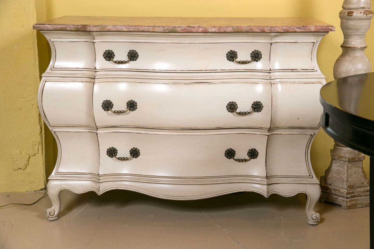 Pair of distress painted marble-top bombe commodes. Magnificent eye-catching beauties. The soft crème/off-white paint will add warmth and character to any place in the home. The antiqued pulls add old world charm to these functional three drawer