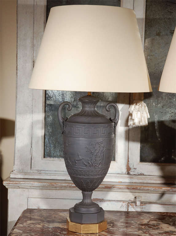 Beautiful pair of Amphora-style Wedgwood type basalt urns turned into lamps. Each featuring a 360 degree, neoclassical band of relief figures beneath a Greek Key pattern. The whole on octagonal, giltwood feet.