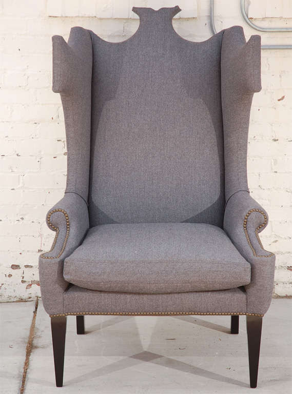 Large wingback chair with unique silhouette, grey wool upholstery, black lacquered legs, and nailhead detailing.