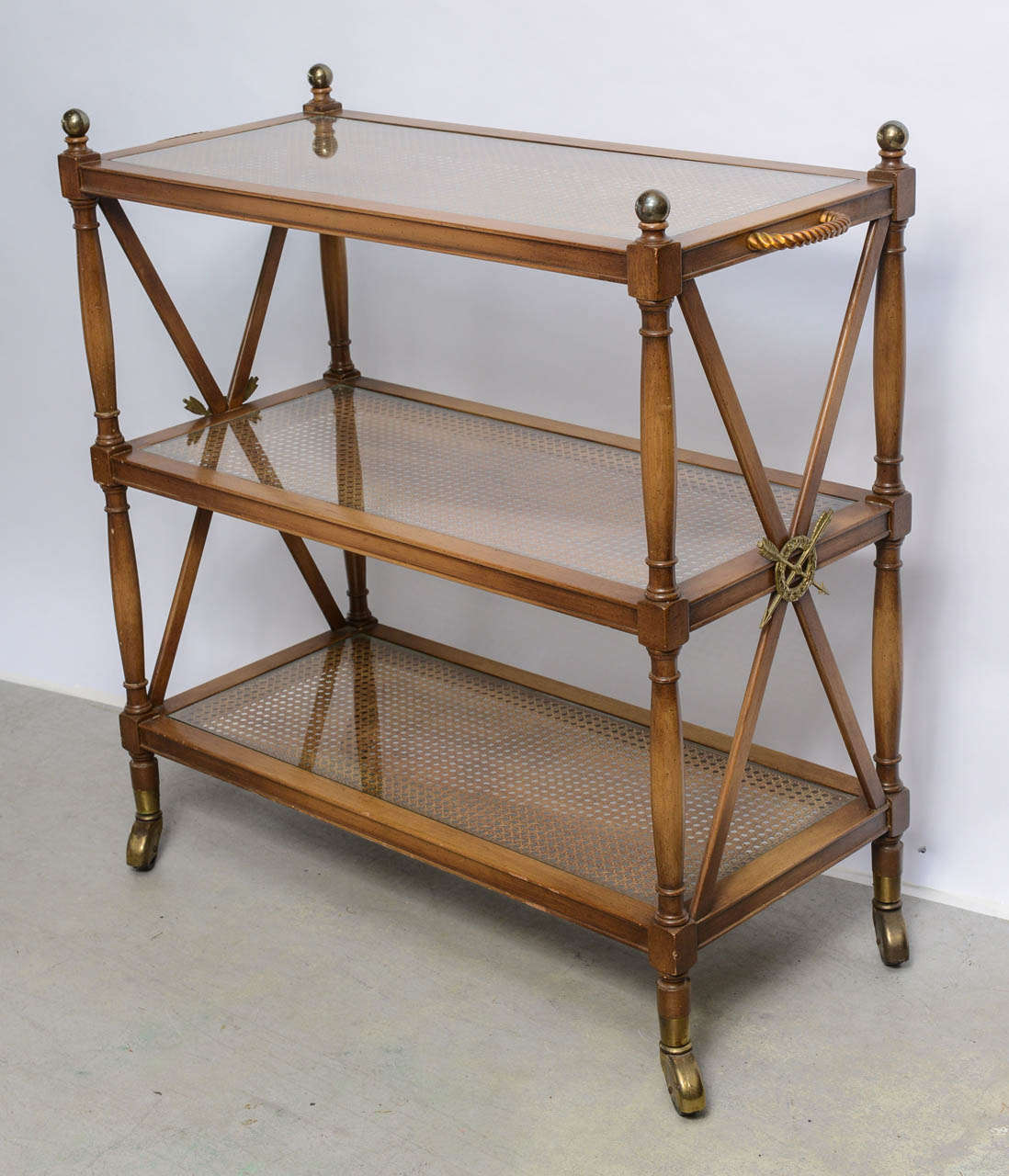 Classic three-tiered trolley with glass shelves inset over cane. The top shelf is finished with four brass finials and the sides are crossed and adorned with arrows and wreath motif.