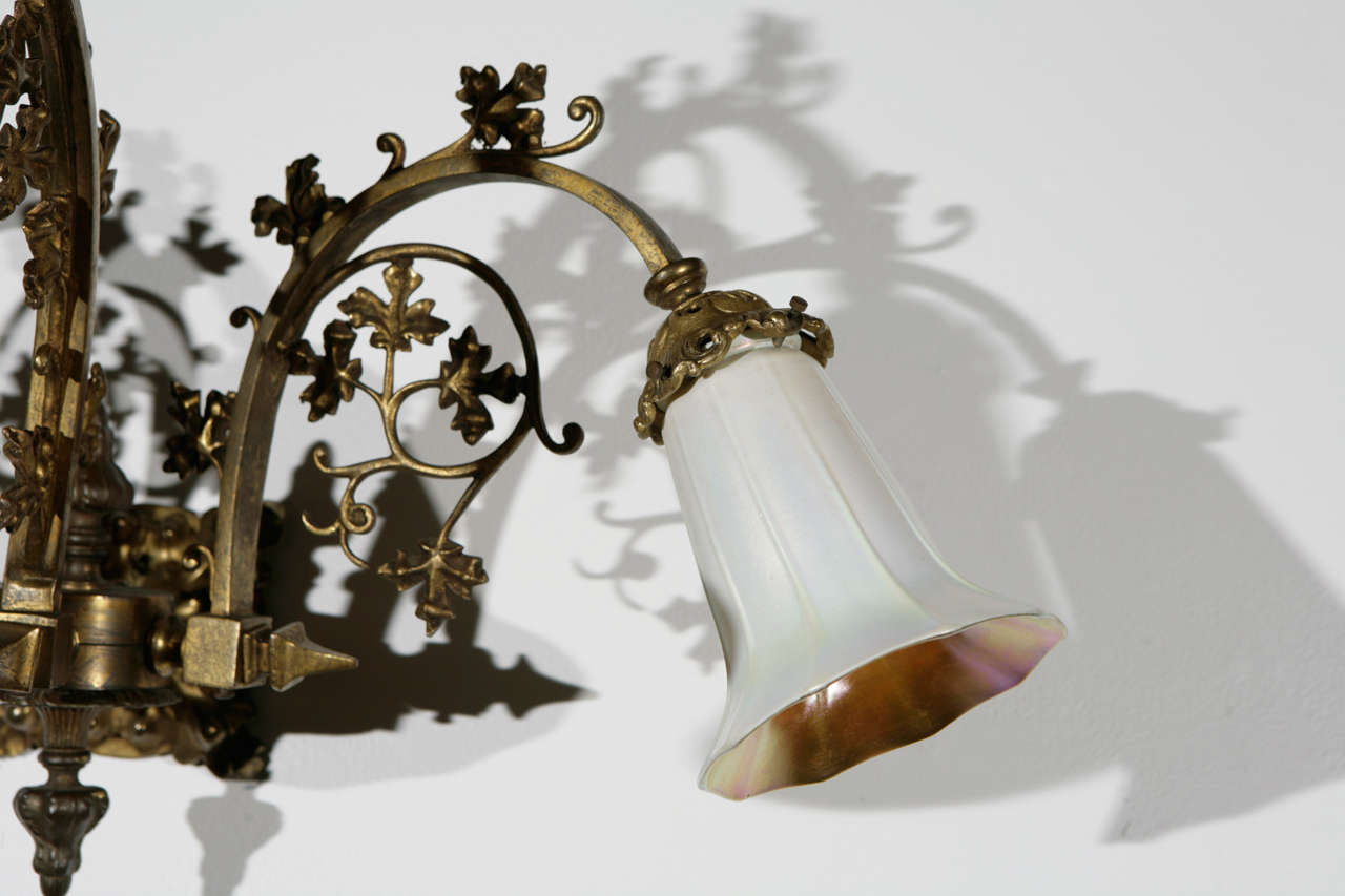 Three-arm cast brass Gothic sconce with signed Quezal glass shades. The shades are not original to the fixture, but are of the same period as the fixture, and are appropriate for the fixture.