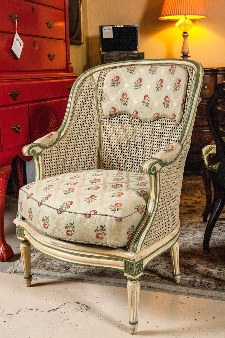 Pair of bergere chairs in the style of Louis XVI. A soft crème fabric with a soft pinkish/red rose floral covers down cushions and back panel. Paint decorated with a crème color designed for a distressed look. The trim of the arms and the leg