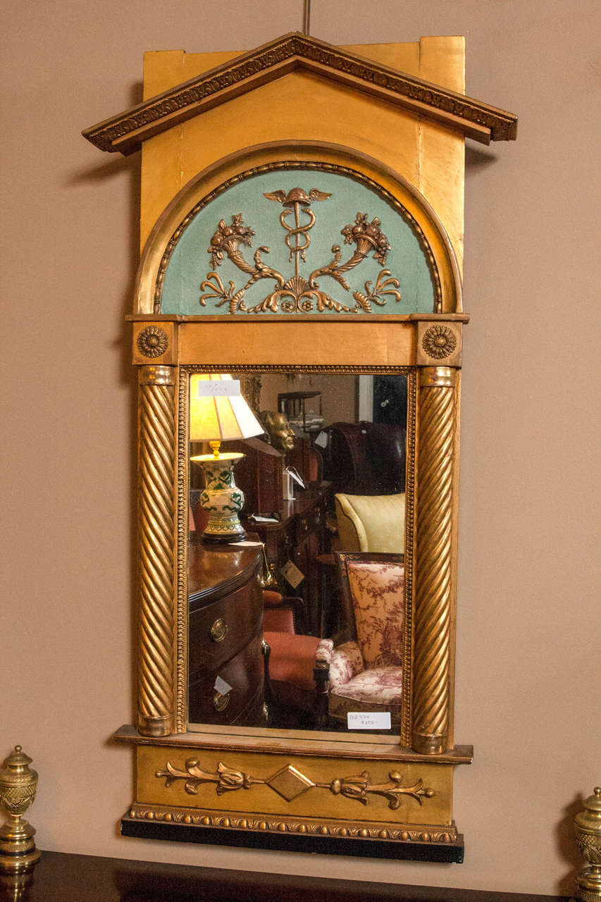 Italian neoclassical pier mirror adorning a caduceus amongst branches topped with flowering baskets along with roots and vines intertwined at the bottom. The background is a soft teal. The mirror's sides have classical wooden columns and medallions.