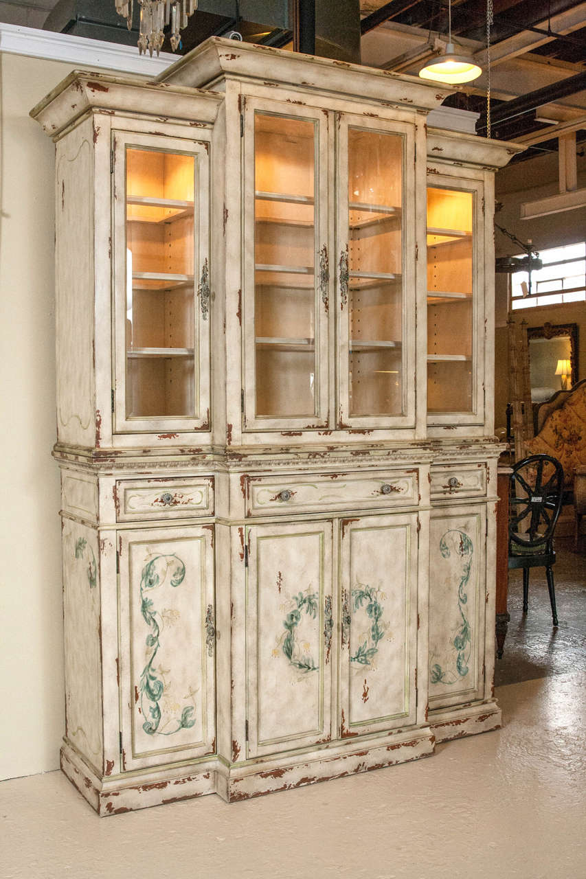 Two-piece paint decorated China cabinet or breakfront. In the style of Adams - this wonderful piece has a lighted interior adding a warm golden hue to whatever one wishes to display. A distressed crème color with a sea blue floral design.