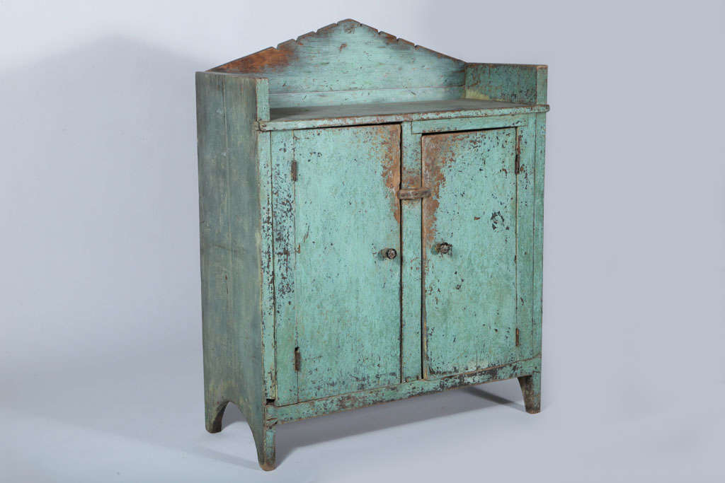 Solid Two-door cupboard with two shelves and primitive door lock. Cut-out style legs and detailed a-frame top. Beautiful turquoise patina exposing weathered paint.
