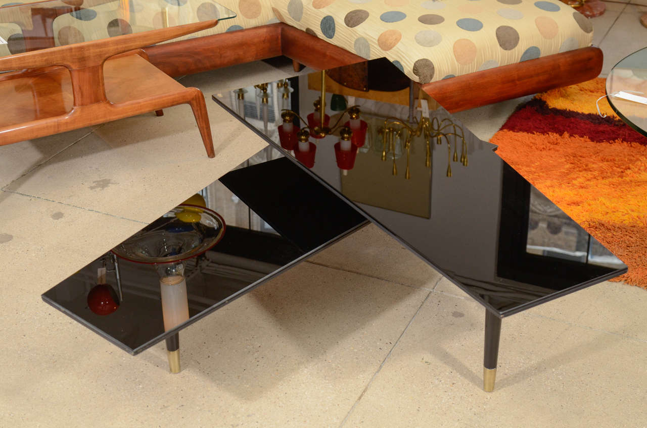 Arrestingly elegant mid-century bi-level multi-directional cocktail table.  Asymmetric trapezoidal tops of black mirrored glass applied to wood with tapered edges.  Three plus one tapered black lacquered legs with brass sabots. Tops pivot on a