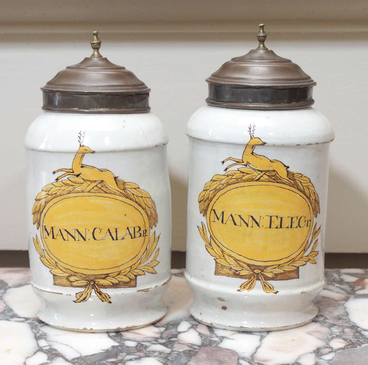 A pair of signed, ceramic tin glazed apothecary jars, with a white ground and an ochre garland encircled cartouche mounted with a stag.  Jars with brass covers.  Jars with some chips to the rim, and wear to the tops.