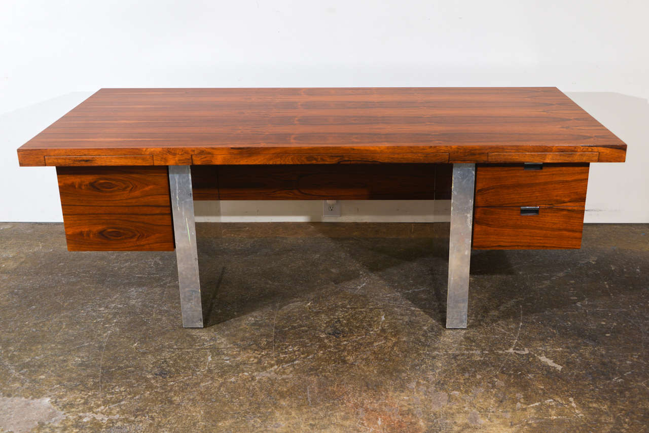 A mid century modern executive desk designed by Roger Sprunger for Dunbar. The desk is made from Rosewood with double chrome pedestal bases. The desk has a set of five drawers including one panel door. We also have a matching credenza available on