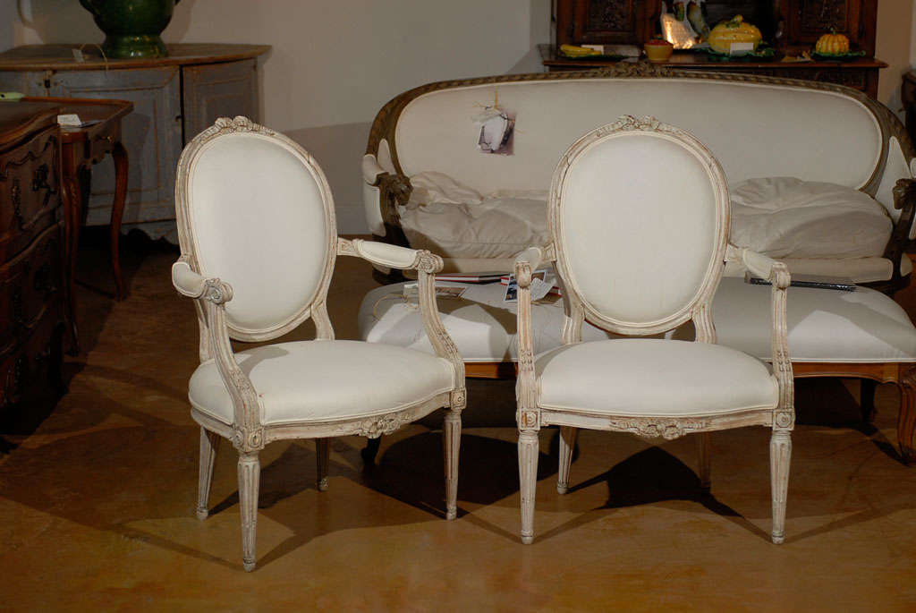 A pair of French Louis XVI style painted wood armchairs from the 19th century with oval backs, scrolled open arms, fluted legs and new upholstery. Each of this pair of French fauteuils features an oval back adorned with a delicately carved ribbon in