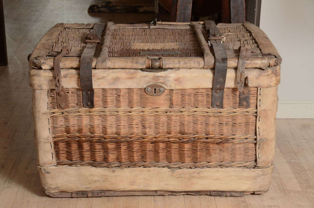 A large heavy wicker basket with leather straps, metal hinges. The edges of the basket are wrapped in waxed goatskin.