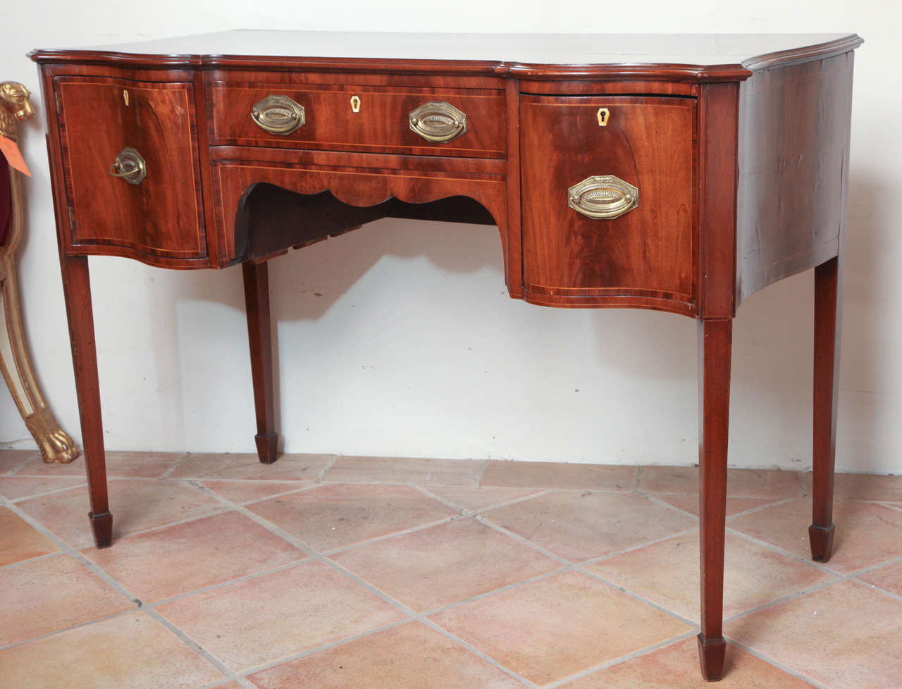 A George III mahogany small-scale serpentine front sideboard fitted with a long central drawer flanked by a cabinet and deep bottle drawer and with crossbanding and stringing throughout.