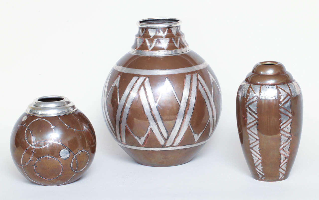 Copper with original bronze color patina and with silver dinanderie geometric designs.

Described from tallest to shortest:
1. 13” high; 10 ¾” wide.
Spherical body with tiered neck and with silver dinanderie geometric linear and V
