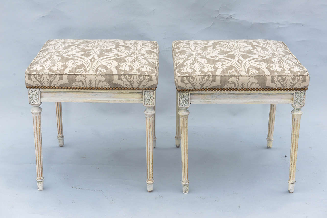Pair of stools, in the Louis XVI style, its painted frame with natural wear, each having a square padded seat, on fielded apron with corner rosettes, raised on round tapering fluted legs, ending in touipe feet. Upholstered in damask printed linen