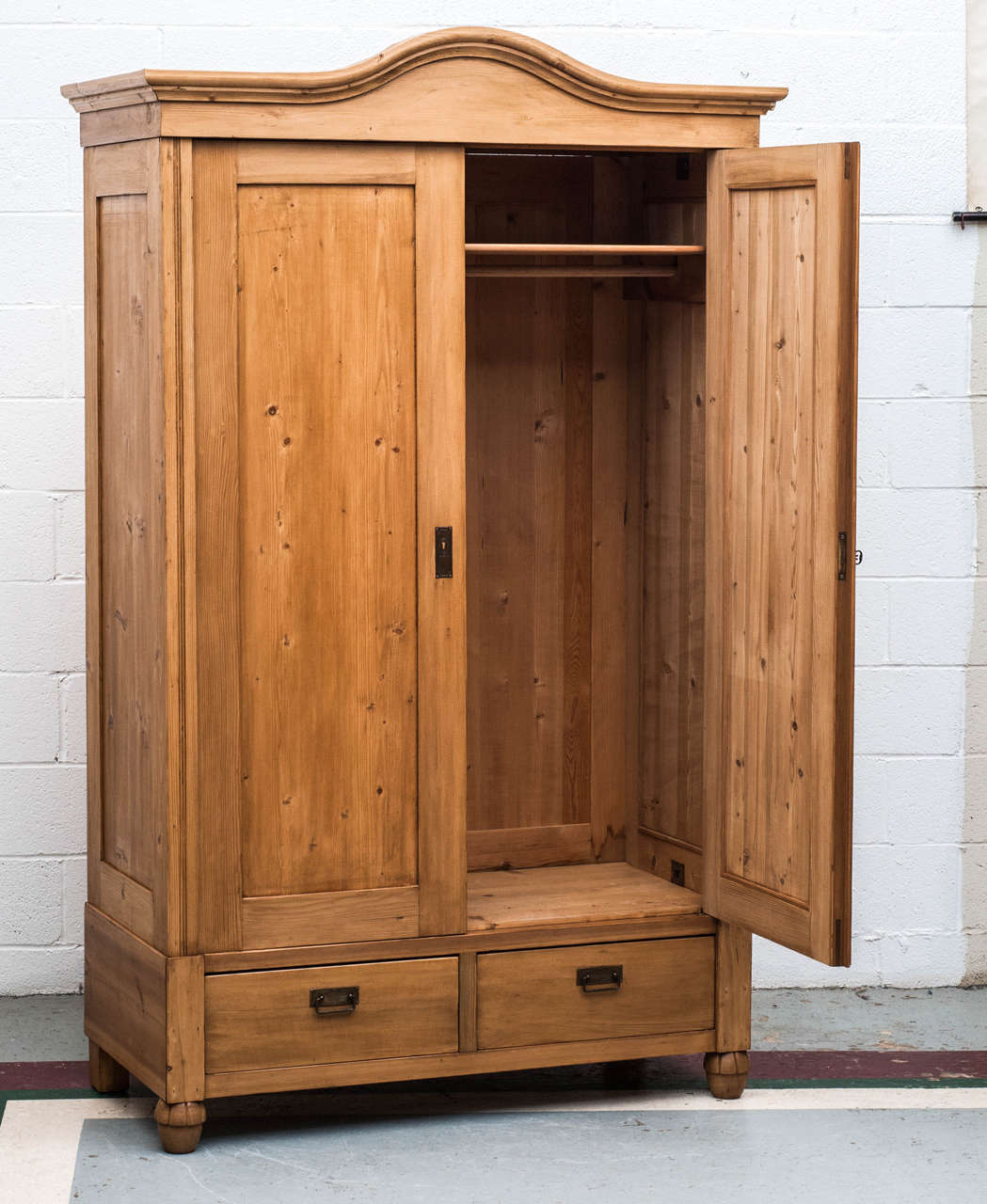 A handsome and spacious bonnet top pine armoire featuring two flat panelled doors, panelled sides and two deep hand-cut dovetailed drawers. Interior has hat shelf and hanging pole. This piece entirely breaks down for easy delivery and access to