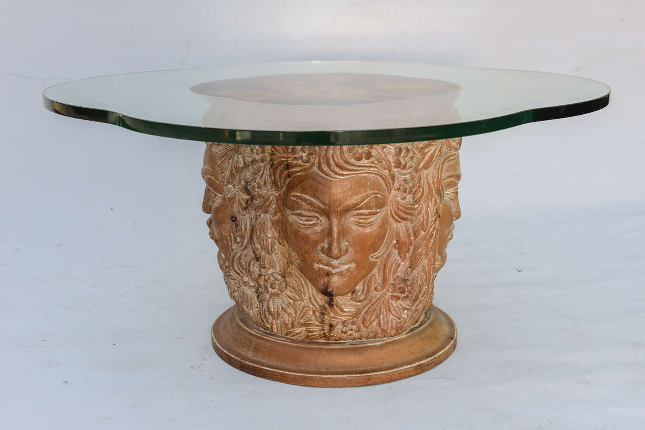 Cocktail table, having a free-form top of glass, centered by tortoise motif eglomise, raised on pedestal base, of pine, each side carved as face relating to a season, on round foot.