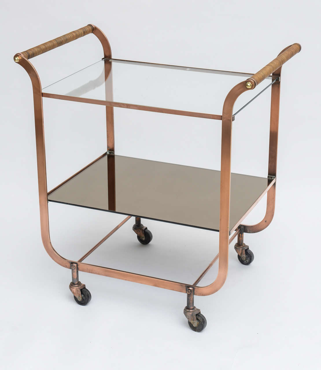 Unusual and handsome Deco-style copper bar cart with rattan-wrapped handles and brass detailing. Top shelf is clear glass, bottom shelf is bronzed mirror. Original wheels are in good condition, and the cart rolls easily.