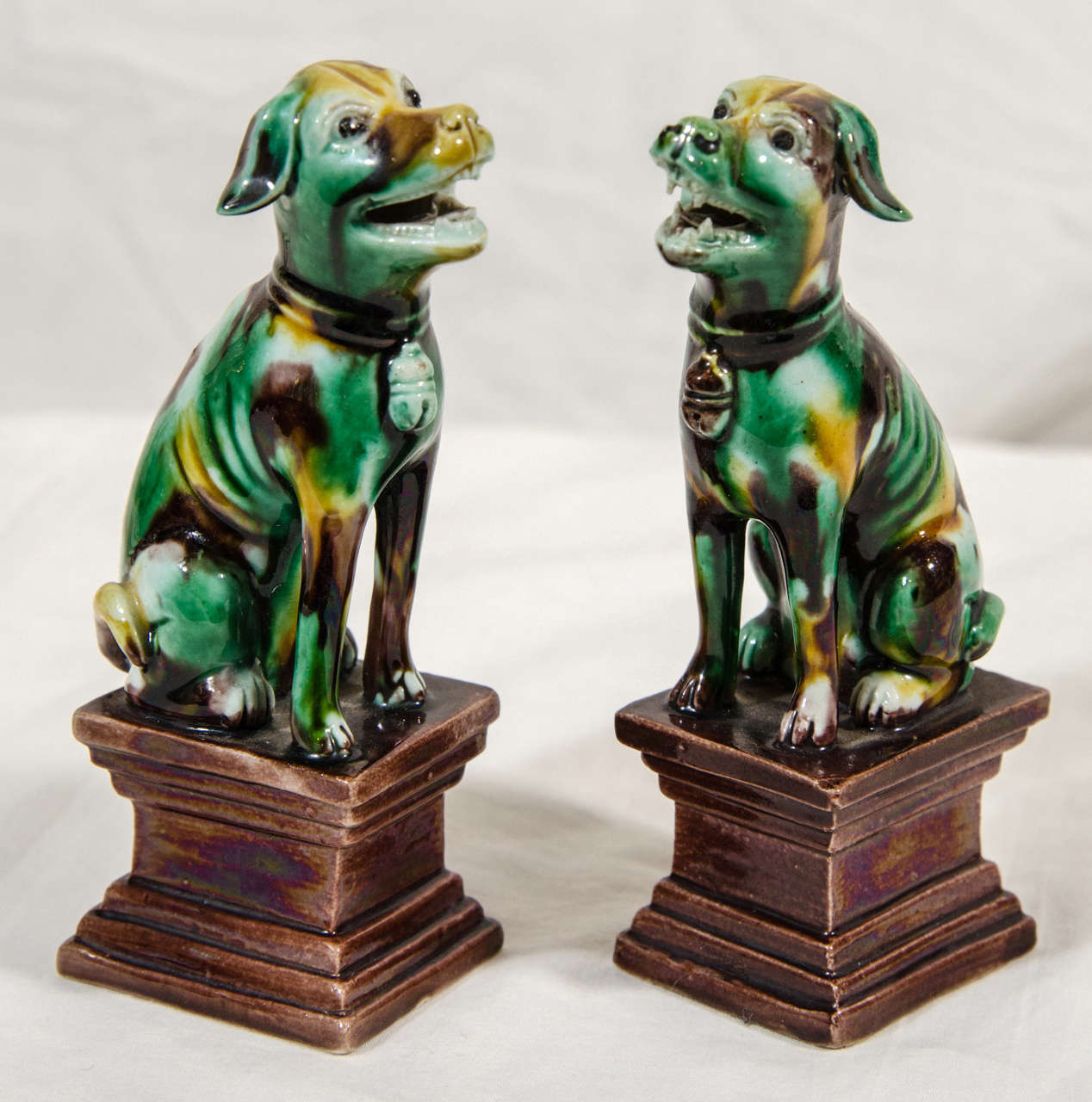 A true pair of Chinese export figures of hounds glazed with 3 color, (Sancai) decoration. Modeled seated on rectangular plinths each turned slightly toward the other. Each with a bell attached to its collar.