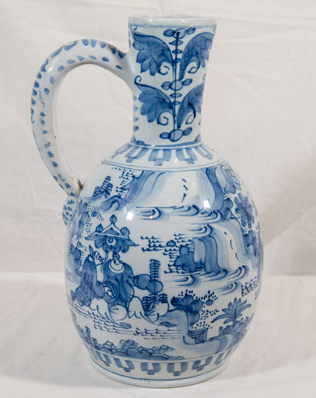 A mid-18th century Frankfurt Delft jug. Decorated all around with chinoiserie garden scenes. Geometric designs incorporating broad flat strokes of dashes and leaves decorate the handle and border.