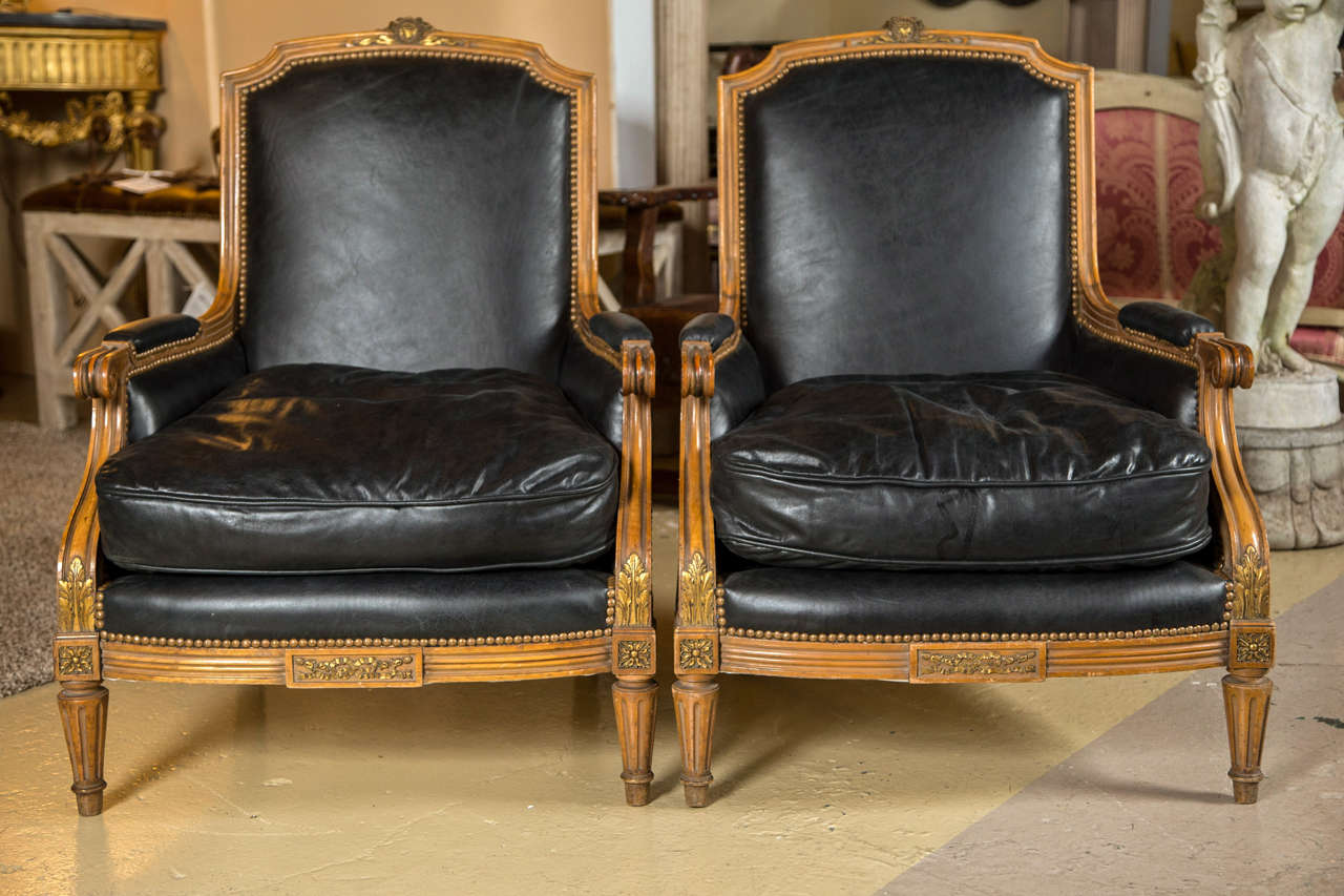 A fine pair of Louis XVI Style French Bergere Chairs possibly by Maison Jansen. The worn black leather upholstery with nail head surround framed with a finely carved and detailed walnut frame. Sides and back also upholstered in worn black leather