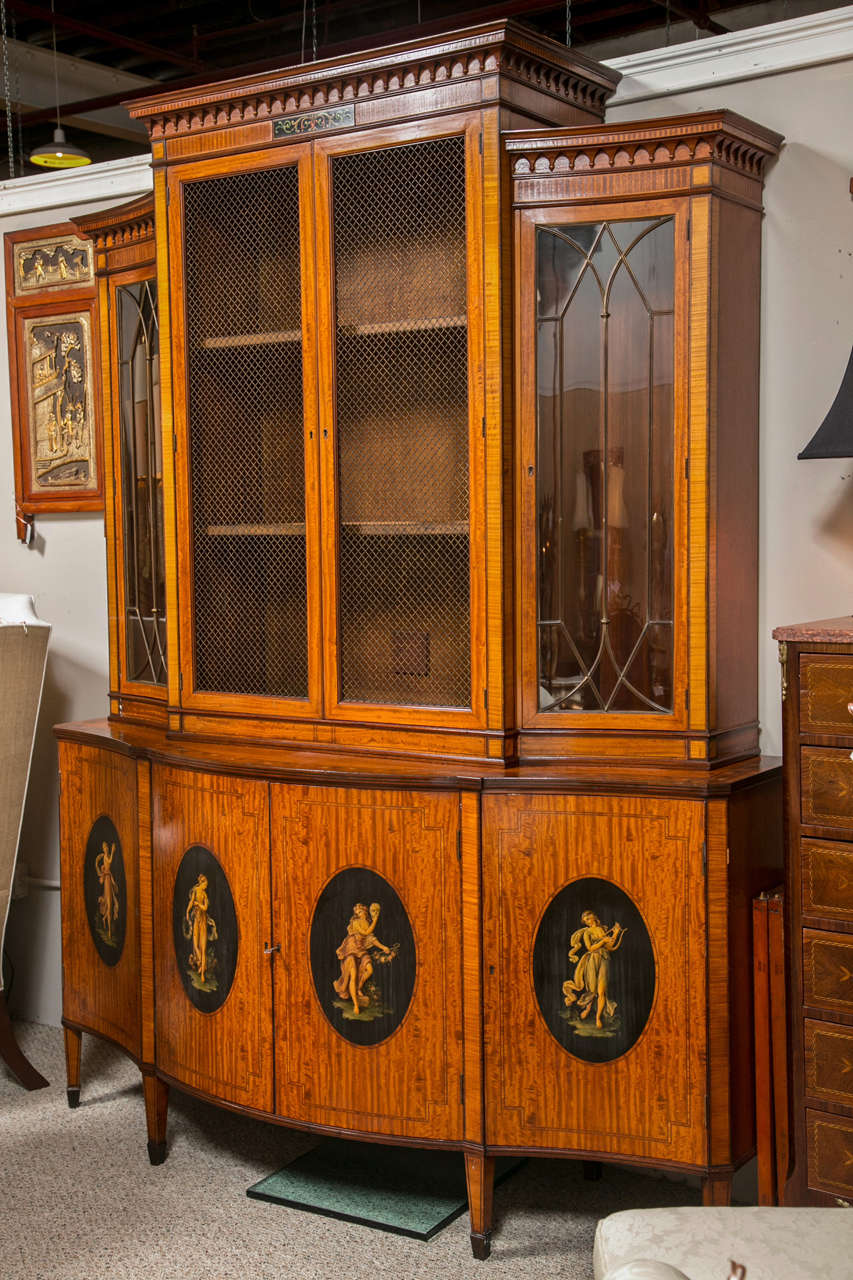 An Edwardian Adams style paint decorated bookcase cabinet. The lower sideboard of satinwood with finely painted lines and Angelica Kauffman style maidens painted inside an ebony oval frame. The upper case with inverted sides and individual glass