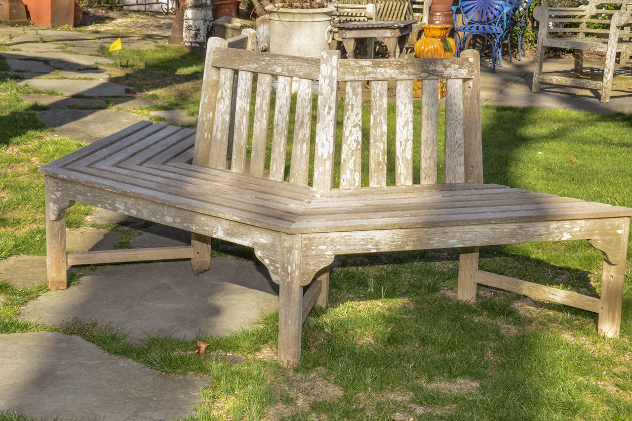 English Teak Tree Surround Bench. The Bench Is Meant To Be Placed Around One Side Of A Tree.It Has A Tapered Slated Vertical Back & Slatted Seat Above 4 Bracketed Square Legs  -- Joined By Stretchers To The Back Legs.
The Teak Has Aged To Beautiful