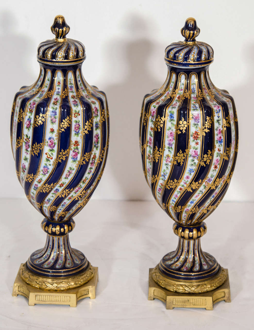 A Pair of Exquisite Antique French Sevres style cobalt porcelain gilt bronze mounted covered urns. Each unique covered vase is hand painted and gilded in great detail depicting multi color flowers and leaves in a swirl pattern finally rested on gilt