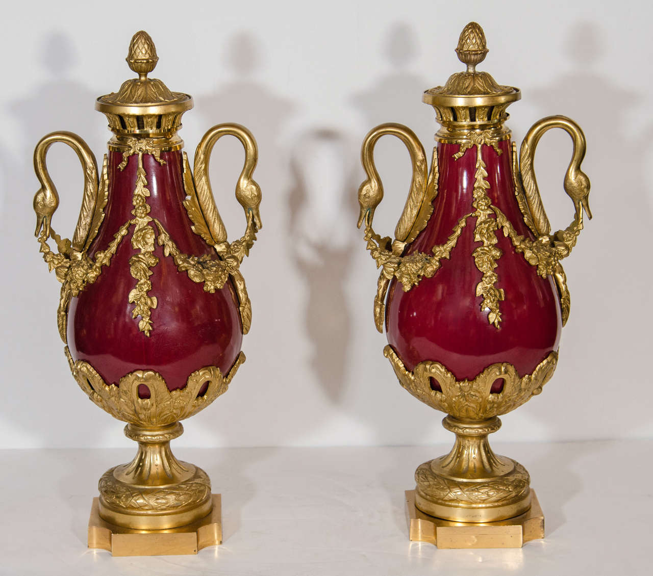 Pair of antique French Louis XVI style gilt bronze and red sevres style porcelain covered urns embellished with gilt bronze swan handles and further adorned with floral wreaths.