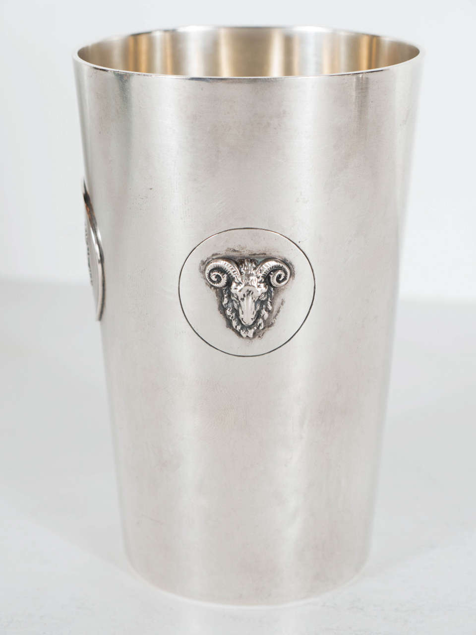 Composed of over six ounces of sterling silver, this beaker features two rams heads in relief surrounded by a chased circle on two sides of the vessel. On a third side there is an image of a rearing horse in relief, circumscribed by a raised circle.