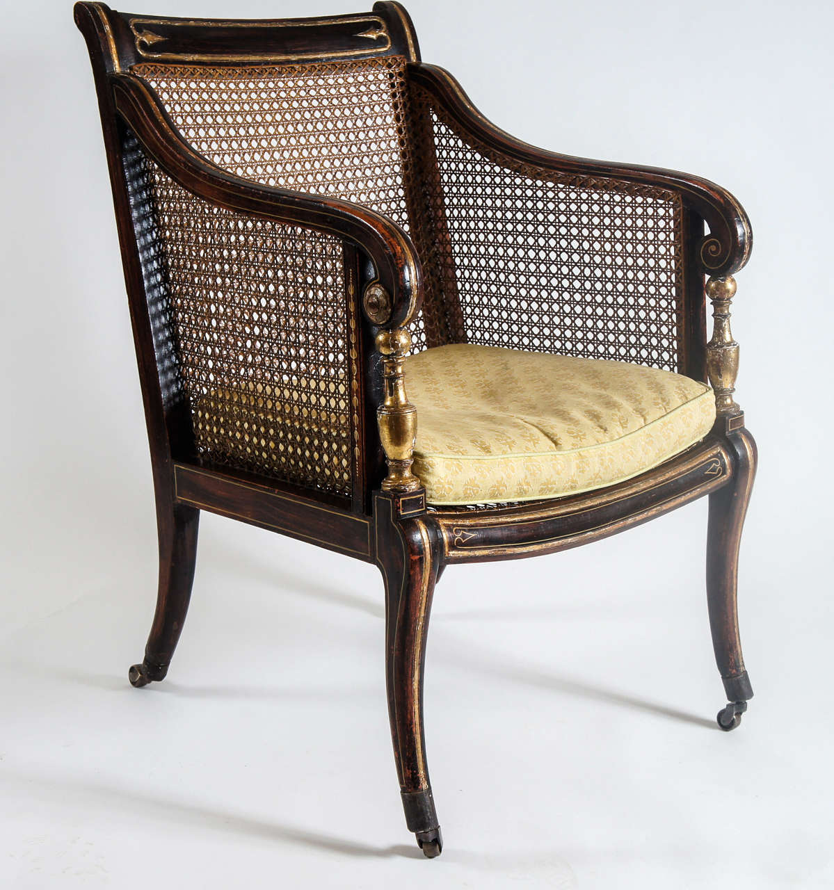 Fine English Georgian Regency period bergere on casters having caned back, sides, and seat with original faux rosewood paint grained frame having parcel gilt carved and painted motifs on top rail, seat rail and legs.  Scrolling arms terminate into