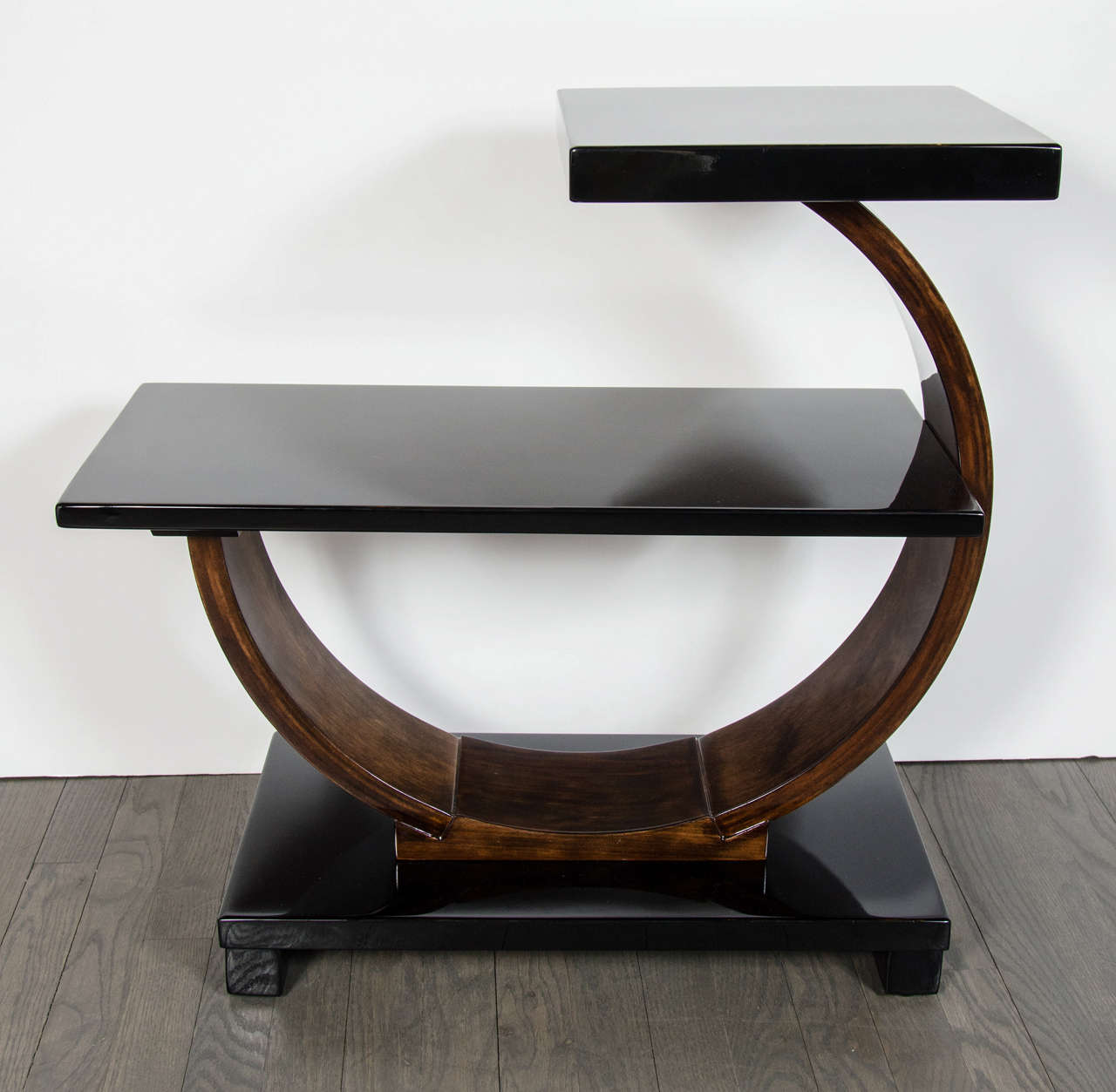 This Art Deco side table by Modernage features a sculptural two tired circular design with a larger lower shelf and smaller top tier supported by an half arc making this piece geometrically interesting and distinctive. It consists of ebonized walnut