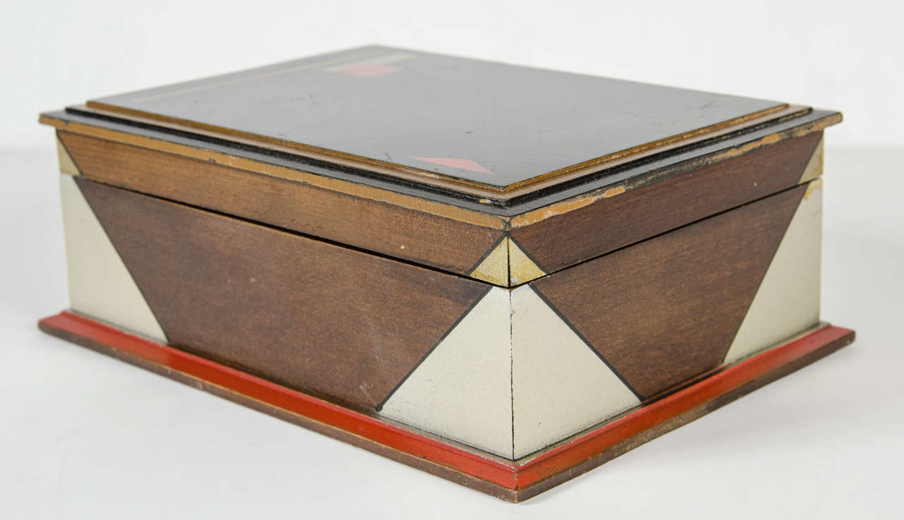 This hand painted box features an abstract Art Deco geometric design with silver leaf,gilt and ebonized detailing as well as hues of platinum and red.