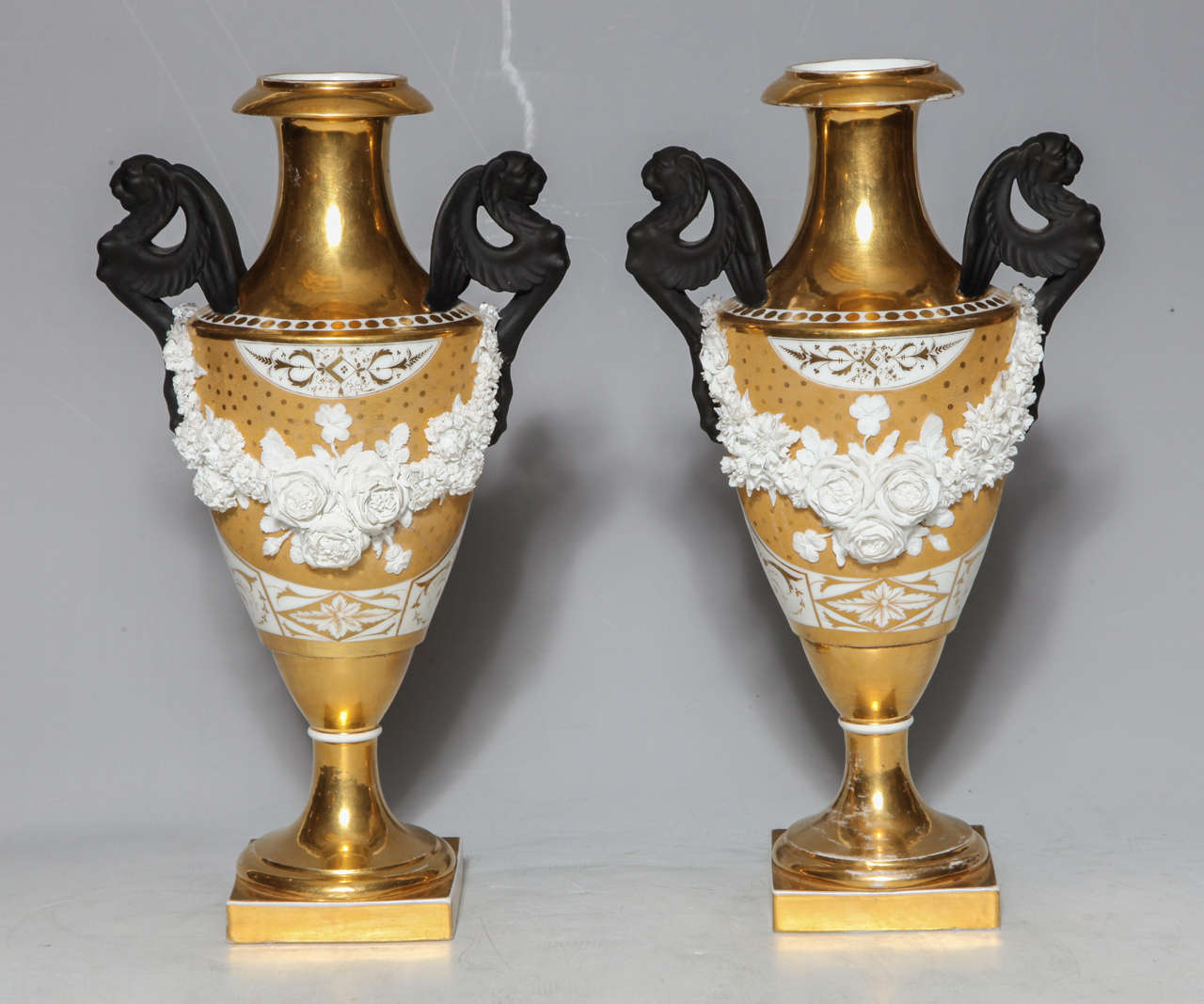 A rare pair of Neoclassical period porcelain vases, possibly Russian from the early 1800s. The gilt porcelain vases are decorated with raised bisque garlands of flowers and porcelain winged dragon handles. Each decorated with matte and burnished 24