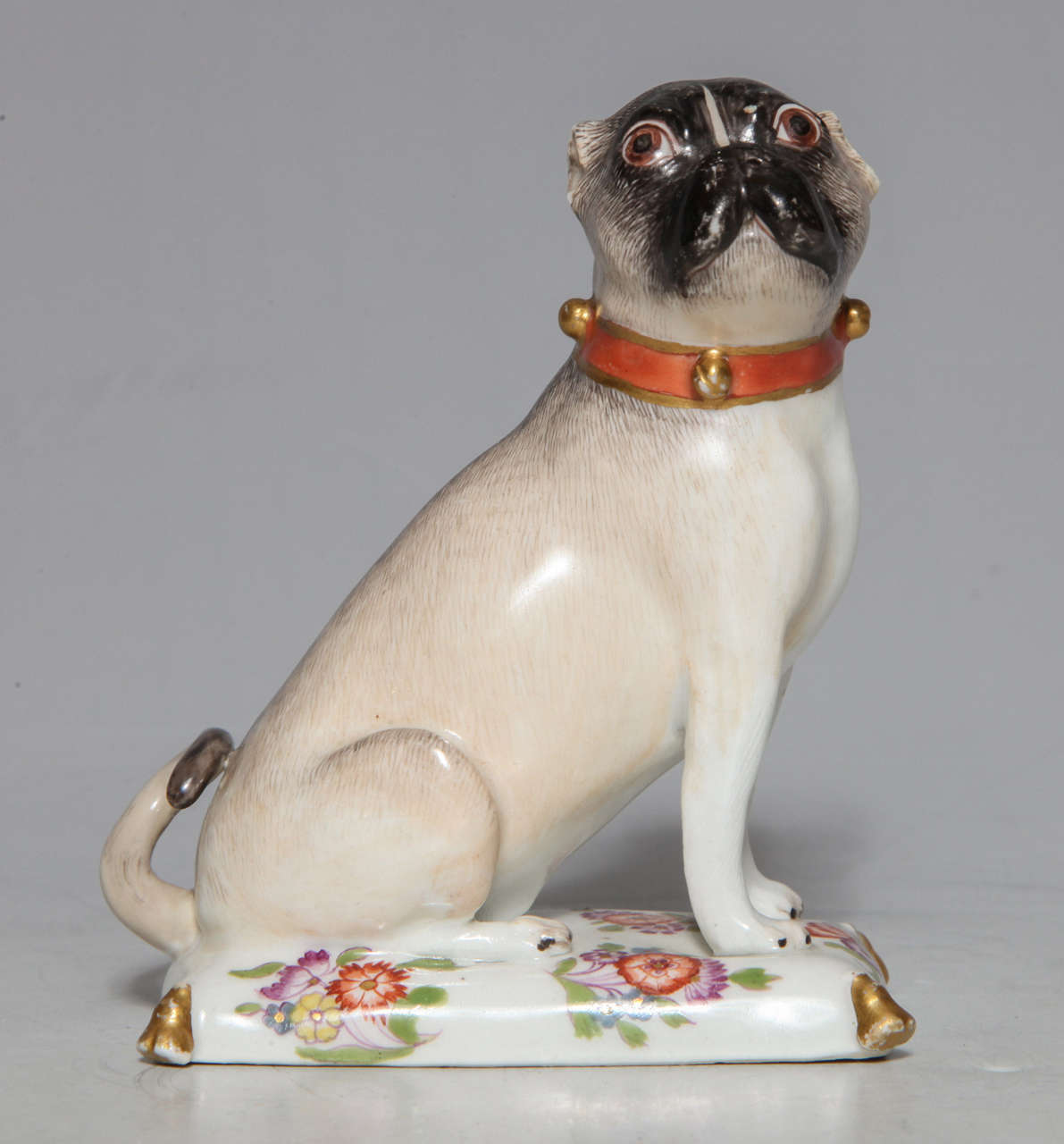18th c. Meissen Porcelain Pug dog seated on a flowered and gold tasseled cushion. Gold colored rings dot the dog's collar with a blue bow positioned at the back of the neck. Molded by J. J. Kandler circa 1750-60.