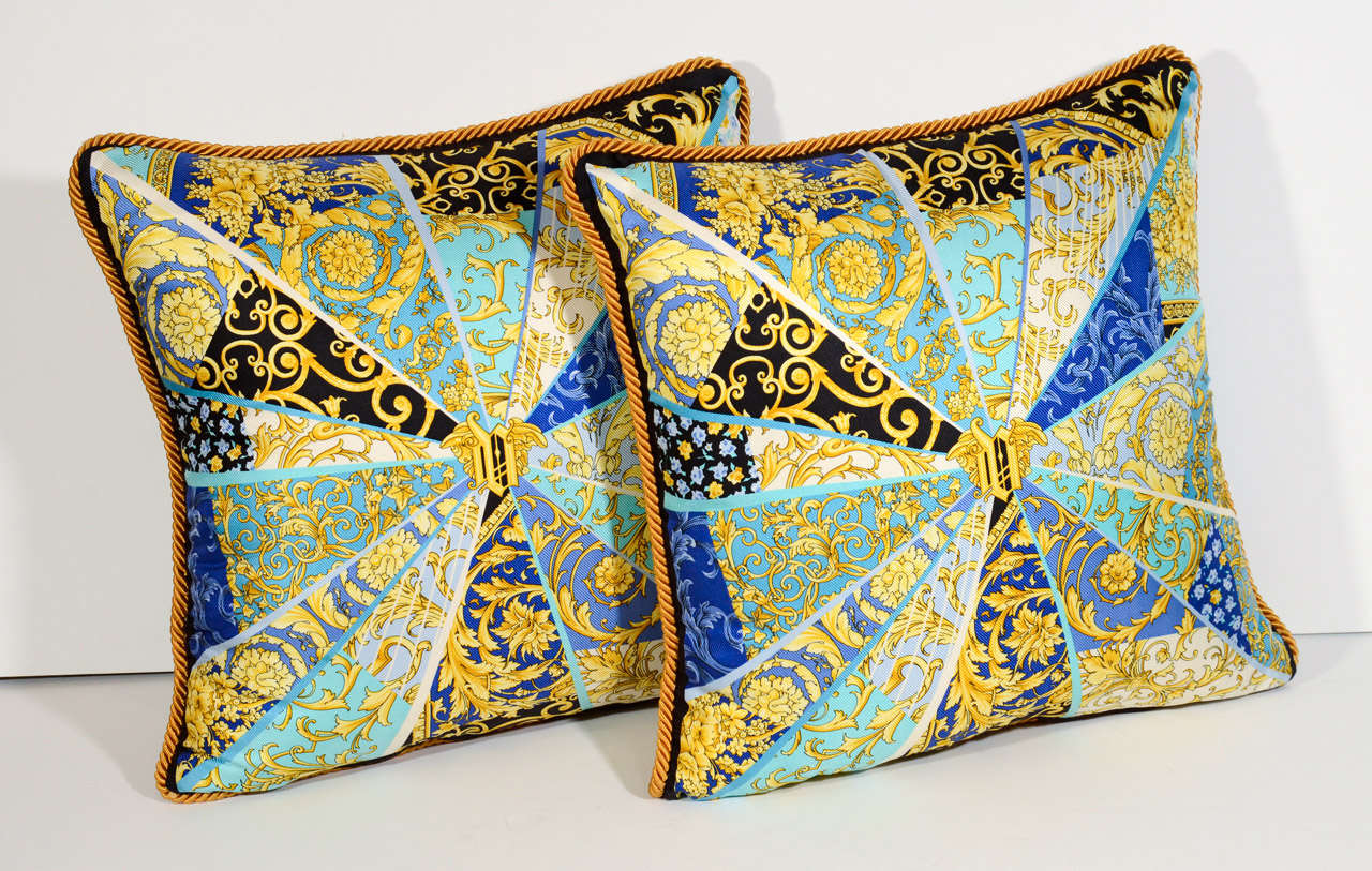 Pair of  Versace decorative pillows with distinguished and renowned scarf prints. The pillows are in silk and feature a variety of vibrant colors and patterns in hues of yellow, blue, turqouise, black, and white.  They have gold braided welting and