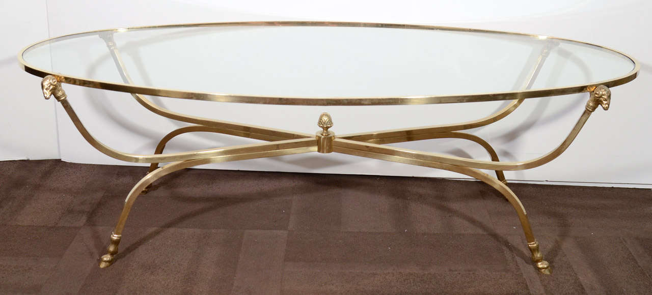 Beautiful cocktail table with oval form comprised of brass frame with stylized rams head and hoofs details, and a glass top. The base has an elegant curved crossed or spider leg design with a pine cone or acorn center detail.  