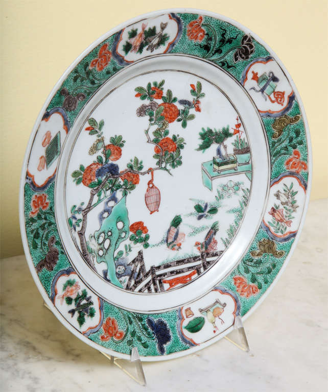 Fine antique Kangxi famille verte circular plate decorated in overglaze enamels, the center with a scholar's garden scene with flowering tree supporting a birdcage containing a gilt bird,rockwork, a bamboo fence and a table set with potted plants