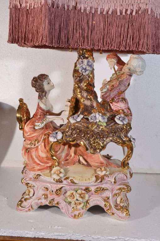 Exquisite, Capo Di Monte Table Lamp, featuring piano playing female and violin playing male with beautiful flowers and exquisite garments in a very large figurine.  Shown with fringed pink custom made lamp shade.  Lamp shade is 16