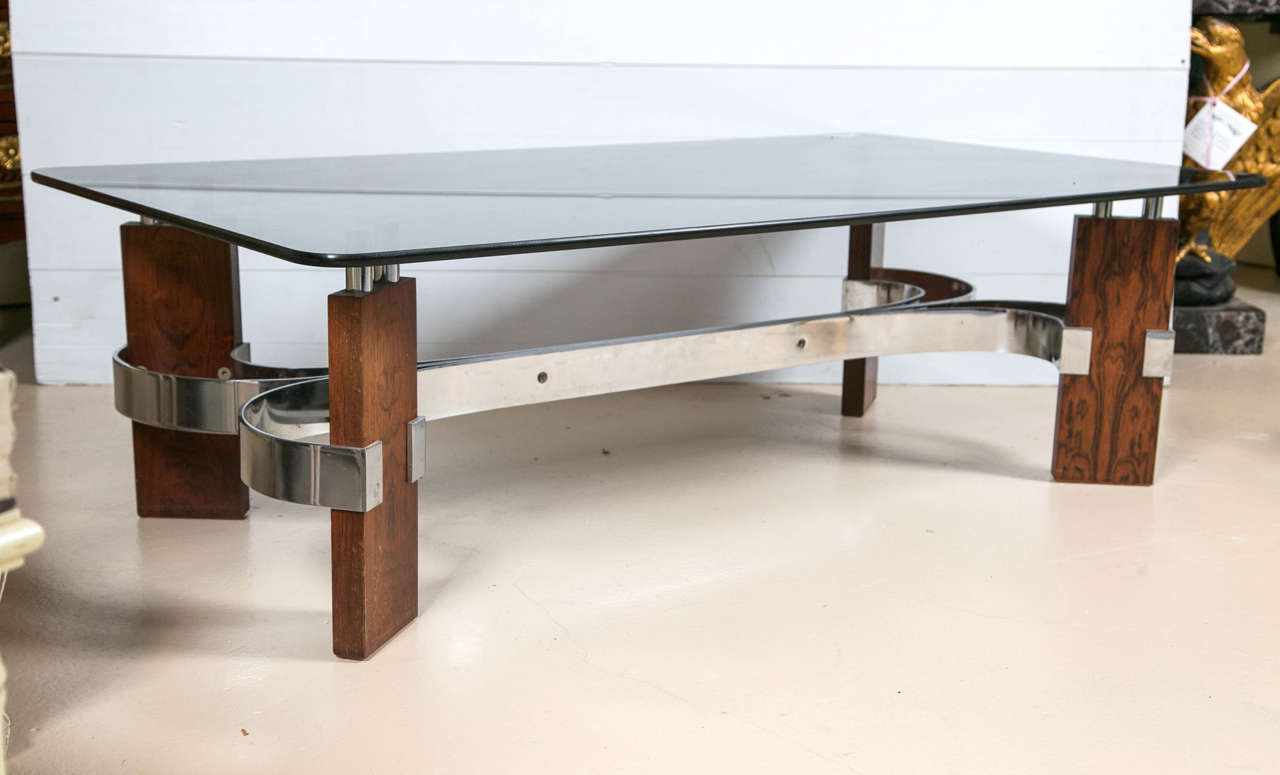 Rosewood and glass top coffee table with a steel stretcher. Mid-Century Modern unique with every curve. The thick wooden legs wrapped with a chrome ‘C‘ grip add to the modern design. A thick smoked glass top sits on two chrome cylinders, great piece.