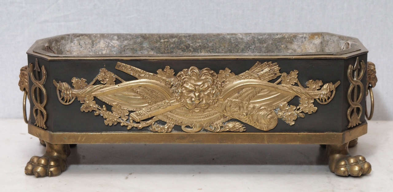 French Empire Patinated and Gilt Bronze Rectangular Footed Jardiniere with ringed Lions head handles and an Empire Cartouche of Assorted weapons and a lions head.