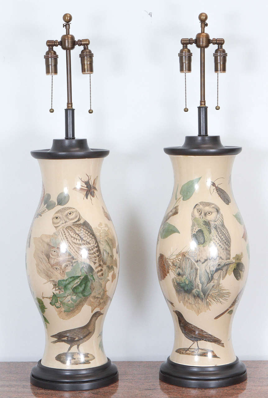Magnificent pair of very large hurricane table lamps. They are decorated with lithographic images of birds and insects, applied to the interior of the glass, with an all-over beige background. The wooden base and cap have a bronze finish and the