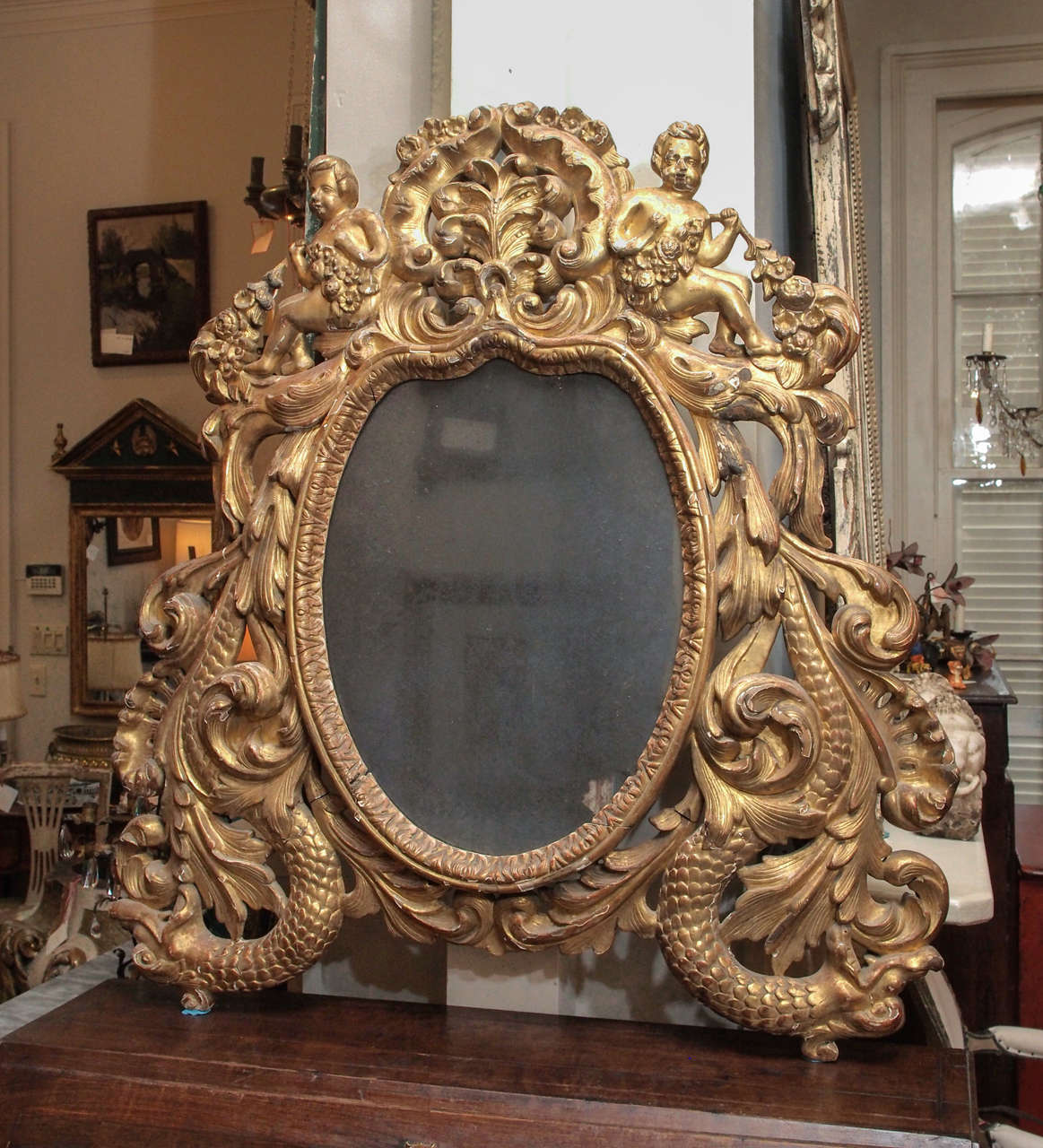 An oval mirror with extravagantly carved frame of sea serpents supporting carved cherubic figures.  Glass original and oxidized.  We've catalogued this piece as Italian, but it might also be from the Iberian peninsula.