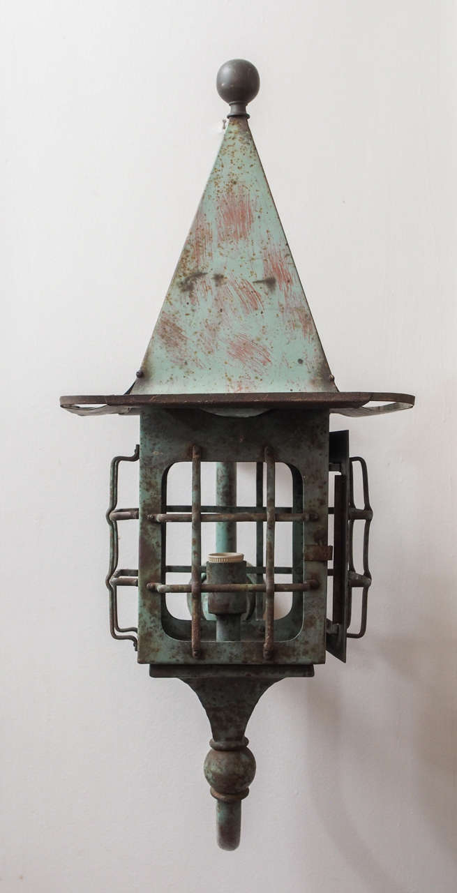A painted wall light, in lantern form, with a tall, pyramidal top with a wide brim, capped with a ball; the lantern openings covered with a grate. Mounted on a U turned pipe and two molded caps.
