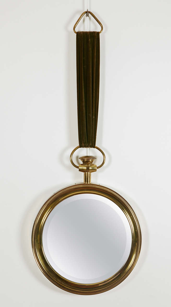 Pocket watch shape mirror by Piero Fornasetti (1913-1988). Brass frame held by a green velvet ribbon, Italy, 1950s.

Total height with ribbon 102cm.
Mirror diameter with frame: 40cm.